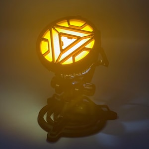 May include: A black and yellow glowing arc reactor replica. The reactor has a circular design with a geometric pattern and is mounted on a black base. The light is bright and casts a warm glow.