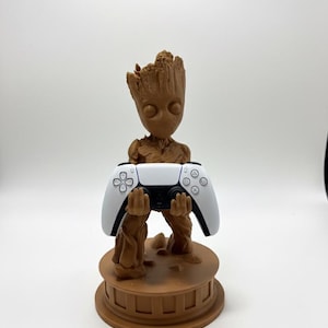 May include: A brown Groot-themed controller stand holding a white and black PlayStation 5 controller. The stand is a detailed sculpture of Groot from Guardians of the Galaxy, with a round base.