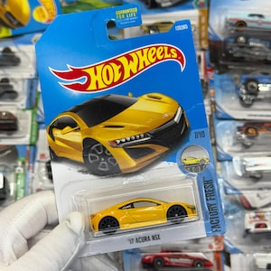 May include: A yellow '17 Acura NSX Hot Wheels die-cast car in a blue and white package. The car is a bright yellow with black wheels and a black roof. The package has the Hot Wheels logo and the words "FACTORY FRESH".