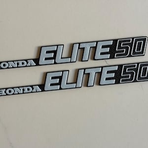 Puede incluir: Dos calcomanías negras y plateadas de Honda Elite 50. La palabra "HONDA" está en plata, seguida de "ELITE 50" en letras plateadas con fondo negro. Las calcomanías están sobre una superficie clara.