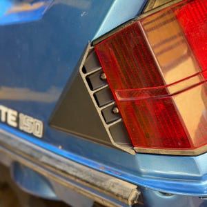 May include: A blue scooter body panel featuring a red rectangular taillight assembly and a black triangular vent insert with visible screws. The partial text "TE 150" is printed in white on the blue surface near the rear light housing.