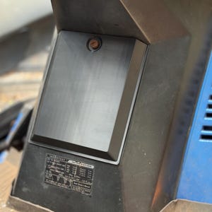 May include: A close-up of a black rectangular panel with a brushed metal finish, featuring a small, round, copper-coloured detail. The panel is part of a larger black structure with a blue, slatted side. A sticker with text is visible.