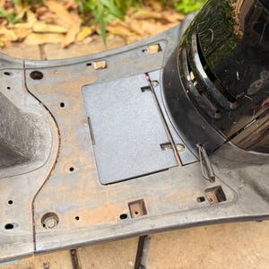 Puede incluir: Vista de cerca del interior de un scooter, que muestra el reposapiés y el compartimento de almacenamiento. La carrocería del scooter es de color gris oscuro con signos de desgaste. Un componente de plástico negro es visible en el lado derecho.
