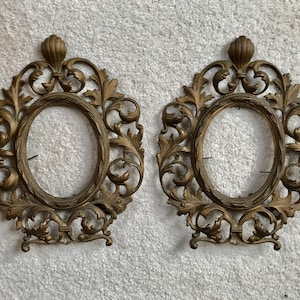 May include: Two ornate gold-toned metal picture frames with intricate scrollwork and oval openings. The frames are empty and ready to display photos or artwork.