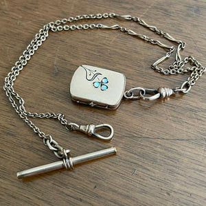 May include: A silver-toned metal watch chain featuring a rectangular locket pendant with a small blue floral inlay design. The chain includes a T-bar toggle clasp at one end and a spring ring clasp, connected by a series of interlocking metal links.