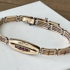May include: A gold-toned metal link bracelet featuring a central elongated oval plaque with three small, round, dark pink stones set horizontally. The bracelet is composed of repeating rectangular segments and includes a secure metal clasp closure.