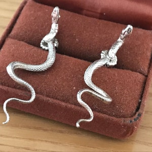 May include: A pair of silver-colored snake earrings. Each earring features a detailed snake design with a coiled body and a raised head. The earrings are displayed in a brown velvet jewelry box.