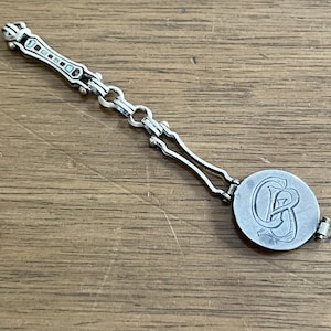 May include: A silver-toned metal watch fob featuring a long, slender bar with small rectangular inlays, connected by a series of circular links to a round medallion. The medallion displays an engraved monogram with the intertwined letters G and B.