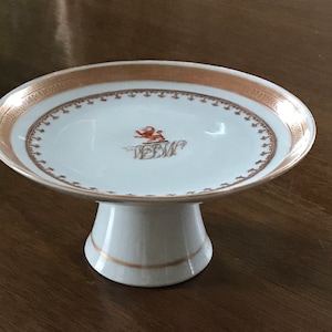 May include: A white porcelain cake stand with a gold rim and a red lion crest with the initials "W.B.W." in the center.
