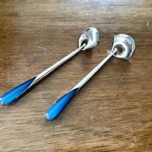 May include: Two silver serving spoons with bright blue enamel accents on the handles. The spoons have a rounded bowl shape and are set against a wooden surface. The spoons are likely for serving food.