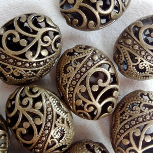 以下が含まれることがあります： 複雑なフィリグリーのデザインが施された、8つの楕円形の金属ビーズのセット。ビーズは濃いブロンズ色で、表面はテクスチャ加工されています。
