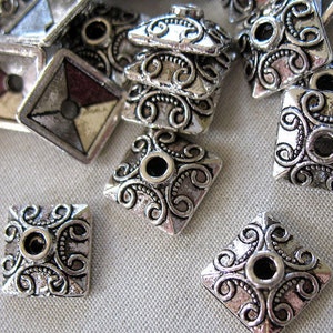 May include: Silver square-shaped metal beads with intricate swirl designs. The beads have a hole in the center for stringing.