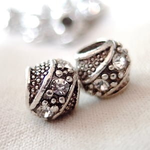 May include: Two silver beads with clear rhinestones. The beads have a textured surface and are designed with a diagonal line of rhinestones.