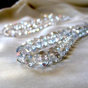 May include: A strand of clear crystal beads, strung together to form a necklace. The beads are faceted and reflect light, creating a shimmering effect.