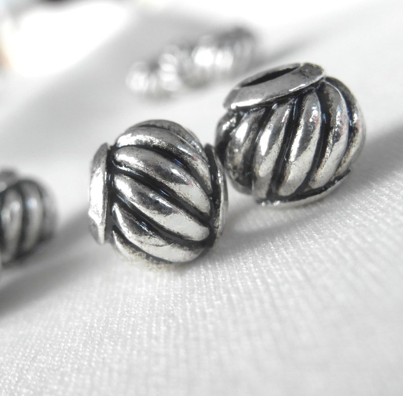 May include: Two silver metal beads with a rope-like design. The beads are round and have a shiny finish.