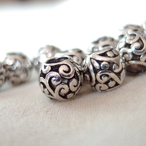 May include: A close-up of a silver charm bracelet with intricate, swirling designs. The charms are round and have a polished finish.