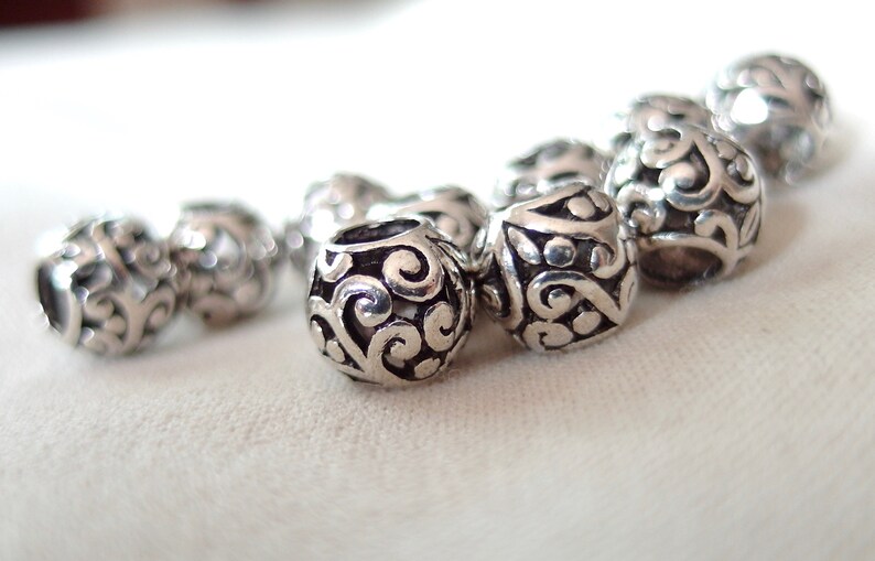 May include: A close-up of a silver charm bracelet with intricate, swirling designs. The charms are round and have a polished finish.