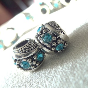 May include: Two silver charm beads with turquoise blue crystal accents. The beads are round and have a textured surface.