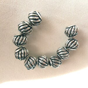 May include: A strand of silver-toned beads with a rope-like design. The beads are arranged in a circle on a white surface.