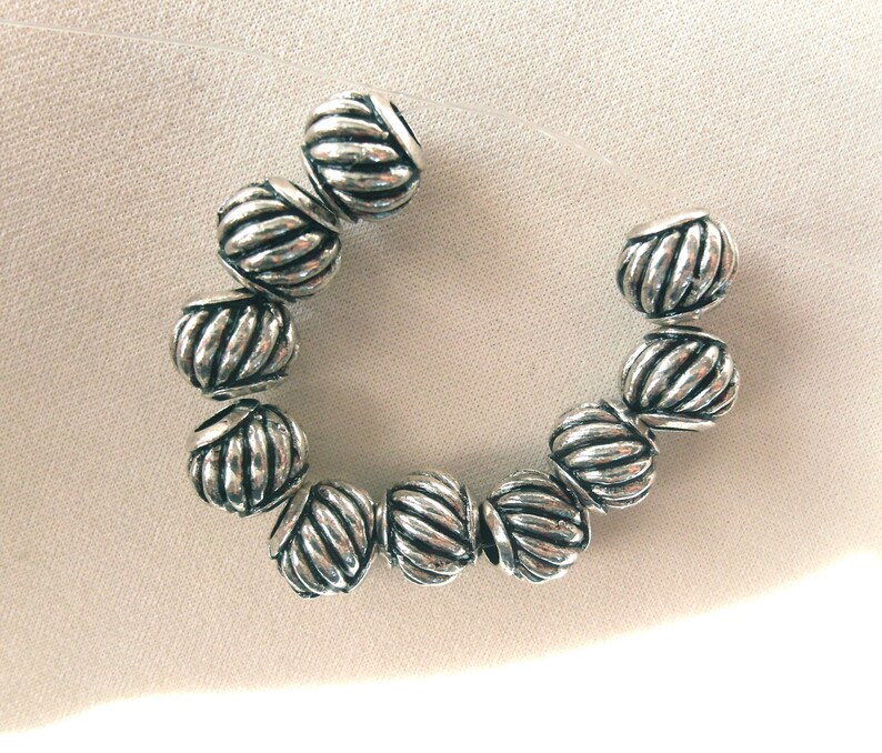 May include: A strand of silver-toned beads with a rope-like design. The beads are arranged in a circle on a white surface.