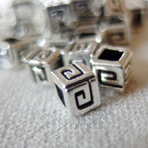 May include: Silver metal beads with a geometric design. The beads are small and have a square shape.