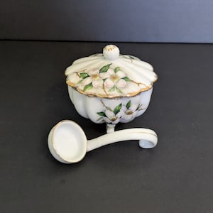 May include: A white ceramic sugar bowl with a lid and a matching spoon. The bowl is decorated with floral patterns in pink, green, and gold. The spoon is approximately 5 inches long.