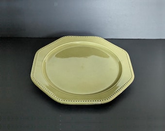 May include: An octagonal ceramic plate in a muted olive green color. The plate features a smooth, glossy finish on the central surface and a raised, textured border decorated with a repeating bead-like pattern along the entire outer edge.
