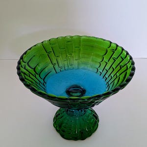 May include: A green glass footed bowl with a basket weave design on the rim and a blue interior. The bowl has a round base and a scalloped edge. The bowl is made of glass and is likely vintage.