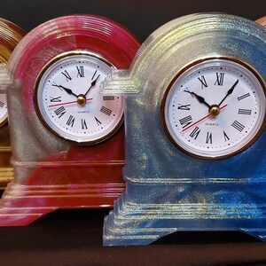 May include: Four decorative quartz clocks with gold-tone, red, and blue resin cases. Each clock has a white face with black Roman numerals, gold hands, and a red second hand. The clocks are on a dark surface.