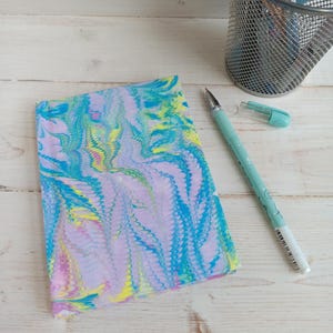 May include: A rectangular notebook featuring a marbled pattern in shades of lavender, bright blue, lime green, and yellow. Beside the notebook lies a light blue ballpoint pen with its cap removed, alongside a silver wire mesh pen holder.
