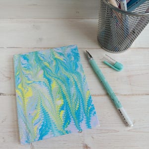 May include: A rectangular notebook featuring a marbled pattern in shades of turquoise, lime green, and white. A light blue ballpoint pen with its cap removed lies next to the notebook, and a silver wire mesh pencil cup holds several pens.