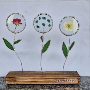 May include: A decorative floral display featuring three glass-encased dried flowers in silver frames. The flowers are mounted on slender metal stems with green glass leaves, set in a wooden base. The display includes a red flower, a cluster of green dots, and a yellow flower.