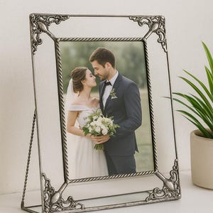 Può includere: Cornice portafoto in metallo argentato con dettagli angolari ornati. Contiene una fotografia di matrimonio. La cornice è esposta su una superficie bianca, con una piccola pianta in vaso sullo sfondo.