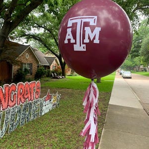 May include: A large maroon balloon with the white letters "A T M" is tied with maroon and white tassels. The balloon is in front of a green lawn with a sign that says "Congrats Audrey 2021".