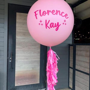 Può includere: Un grande palloncino rosa con il nome "Florence Kay" stampato in rosa, decorato con stelle. Il palloncino è attaccato a una ghirlanda di nappe rosa e a un sottile bastoncino viola. Il palloncino è all'aperto.
