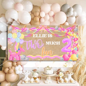 May include: A birthday banner with the text "Ellie is Two Much Fun" in pink and yellow. The banner features a pink balloon unicorn and a large number 2 balloon. The background is a light brown color.