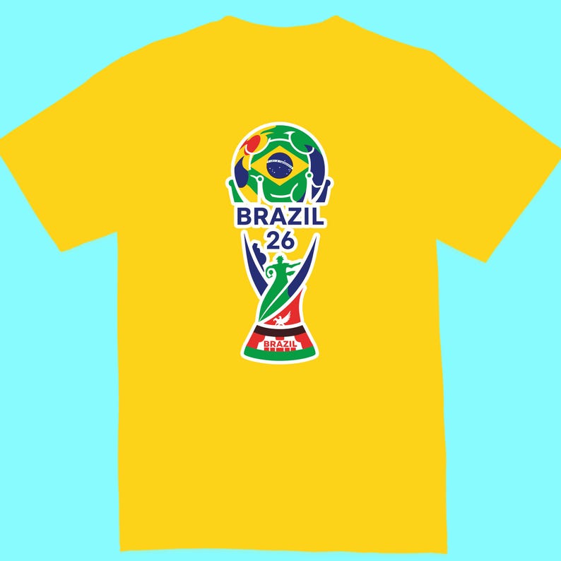 May include: Yellow t-shirt featuring a colorful graphic of a soccer ball and trophy design with the text "BRAZIL 26". The graphic incorporates the colors of the Brazilian flag. The shirt is a short-sleeved crew neck.