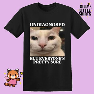 Op de afbeelding: Zwart T-shirt met een close-up van een witte kat met een grijnzende uitdrukking. De tekst "UNDIAGNOSED BUT EVERYONE'S PRETTY SURE" staat boven en onder de kattenafbeelding. Het shirt heeft het "SILLY LITTLE SHIRTS" logo.