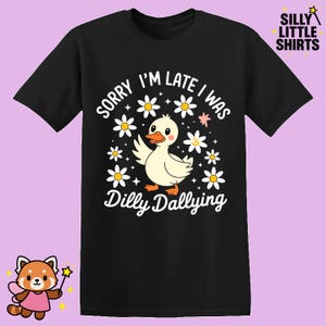 Op de afbeelding: Zwart T-shirt met een afbeelding van een cartoon eend omringd door witte madeliefjes. De tekst luidt "SORRY I'M LATE I WAS Dilly Dallying". Het shirt heeft ook de tekst "SILLY LITTLE SHIRTS" rechtsboven.