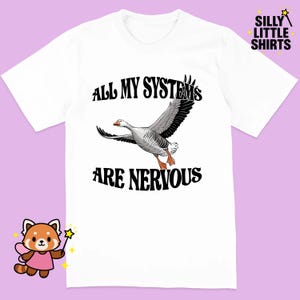 Op de afbeelding: Wit T-shirt met de zwarte tekst "ALL MY SYSTEMS ARE NERVOUS", met een cartoon gans in vlucht. Het shirt heeft ook de tekst "SILLY LITTLE SHIRTS" in de rechterbovenhoek.