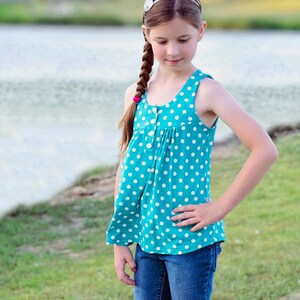 Puede incluir: Una niña pequeña con un top sin mangas azul turquesa con lunares blancos y pantalones vaqueros azules. Lleva una diadema plateada con un pequeño diseño floral. Está de pie en un camino pavimentado con hierba verde y un cuerpo de agua en el fondo.