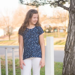 May include: A young person wearing a blue and white patterned short-sleeved shirt and white pants. They are standing next to a white picket fence and a tree.