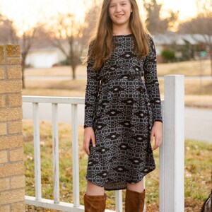May include: A young person wearing a long-sleeved black and white patterned dress with brown knee-high boots. The person is standing in front of a white picket fence.