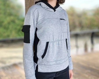 Puede incluir: Una sudadera gris con capucha con detalles negros y un bolsillo en la manga izquierda. La sudadera tiene una cremallera negra y un cordón negro.