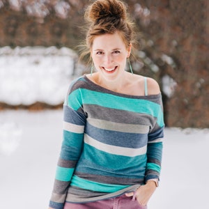 May include: A woman with a messy bun hairstyle is wearing a teal, gray, and white striped long-sleeve shirt with a slightly off-the-shoulder neckline. She is smiling and looking at the camera.