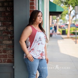May include: A woman wearing a sleeveless top with a floral print and a coral coloured panel. She is wearing blue jeans and brown sandals. The top has a scoop neck and the floral print is in shades of pink, blue, and green.