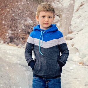 May include: A young boy wearing a blue, white, and gray hooded sweatshirt with a blue drawstring. He is standing on a snowy path and looking to the right.