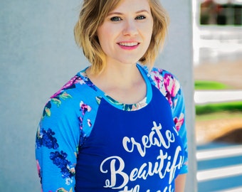 Puede incluir: Una mujer lleva una camiseta de béisbol azul y blanca con el texto "create beautiful things" impreso en la parte delantera. La camiseta tiene un estampado floral en las mangas.