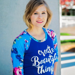 May include: A woman wearing a blue and white baseball tee with the text "create beautiful things" printed on the front. The shirt has a floral pattern on the sleeves.