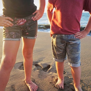 May include: Two children stand on a sandy beach, one wearing floral print shorts and a black shirt, the other wearing camouflage print shorts and a red shirt.